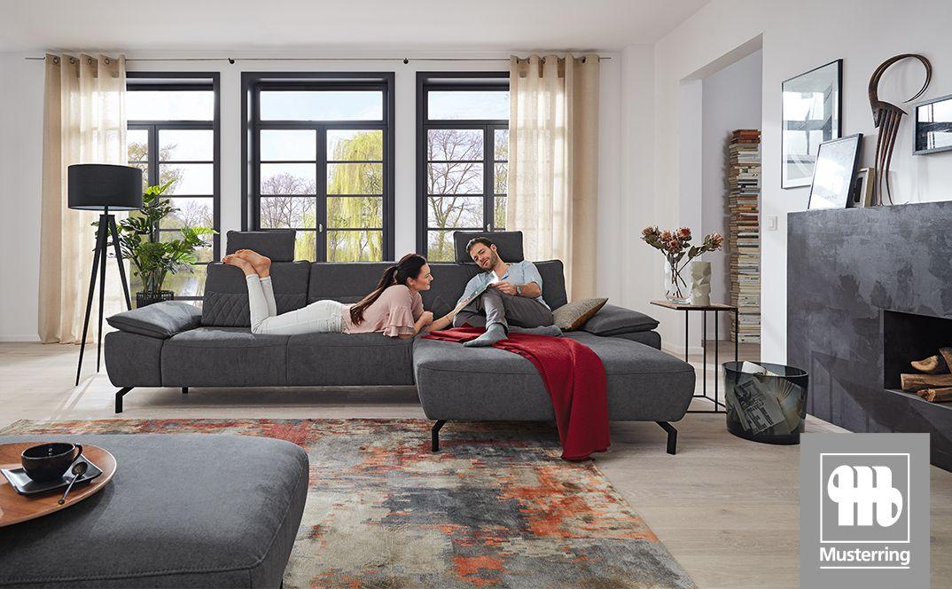 Modernes graues Ecksofa in hellem Wohnzimmer, ein Paar entspannt darauf vor großen Fenstern.