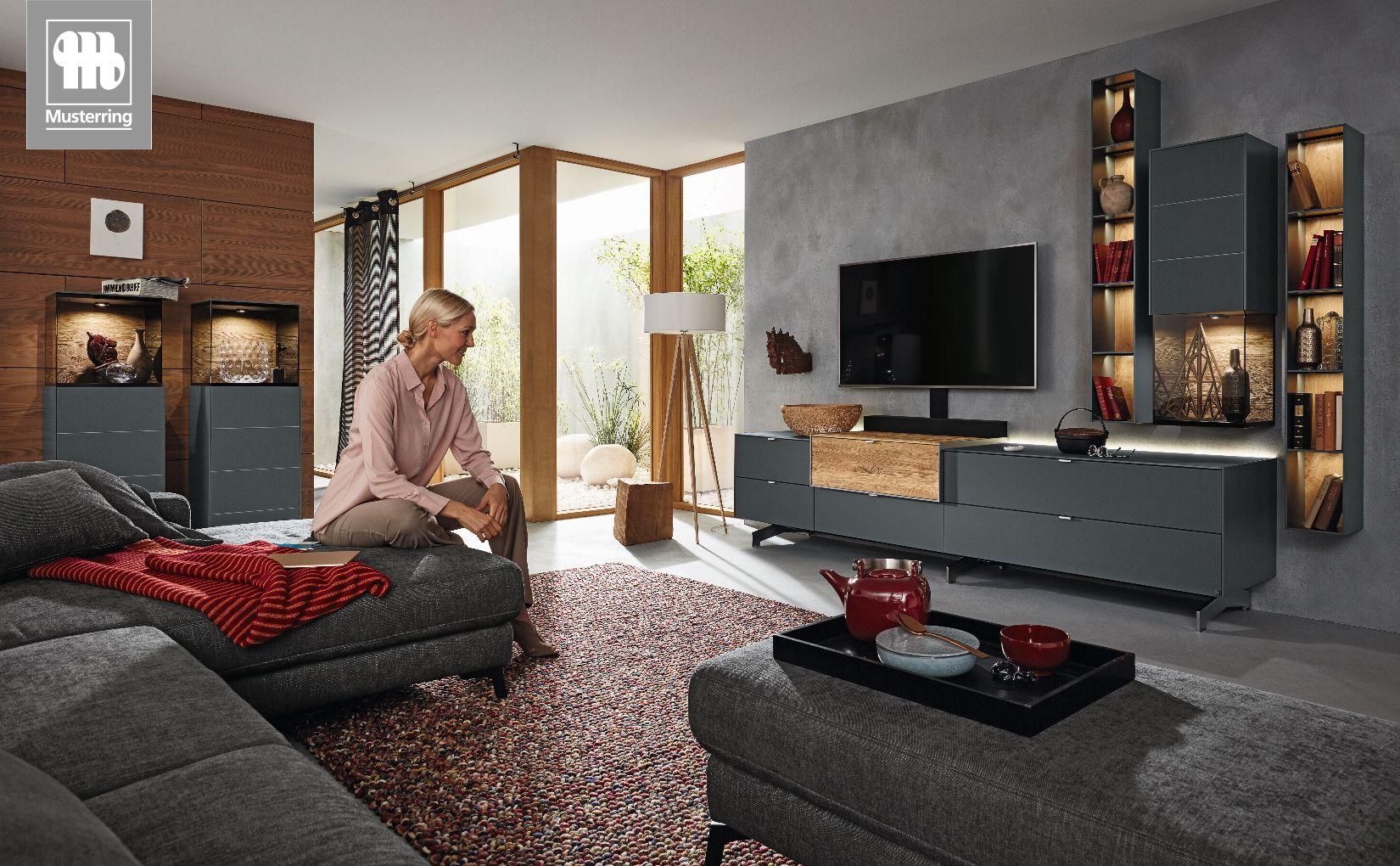 Wohnwand Q-Media 2.0 von Musterring in dunklem Grau mit Holzakzenten und beleuchteten Vitrinen.