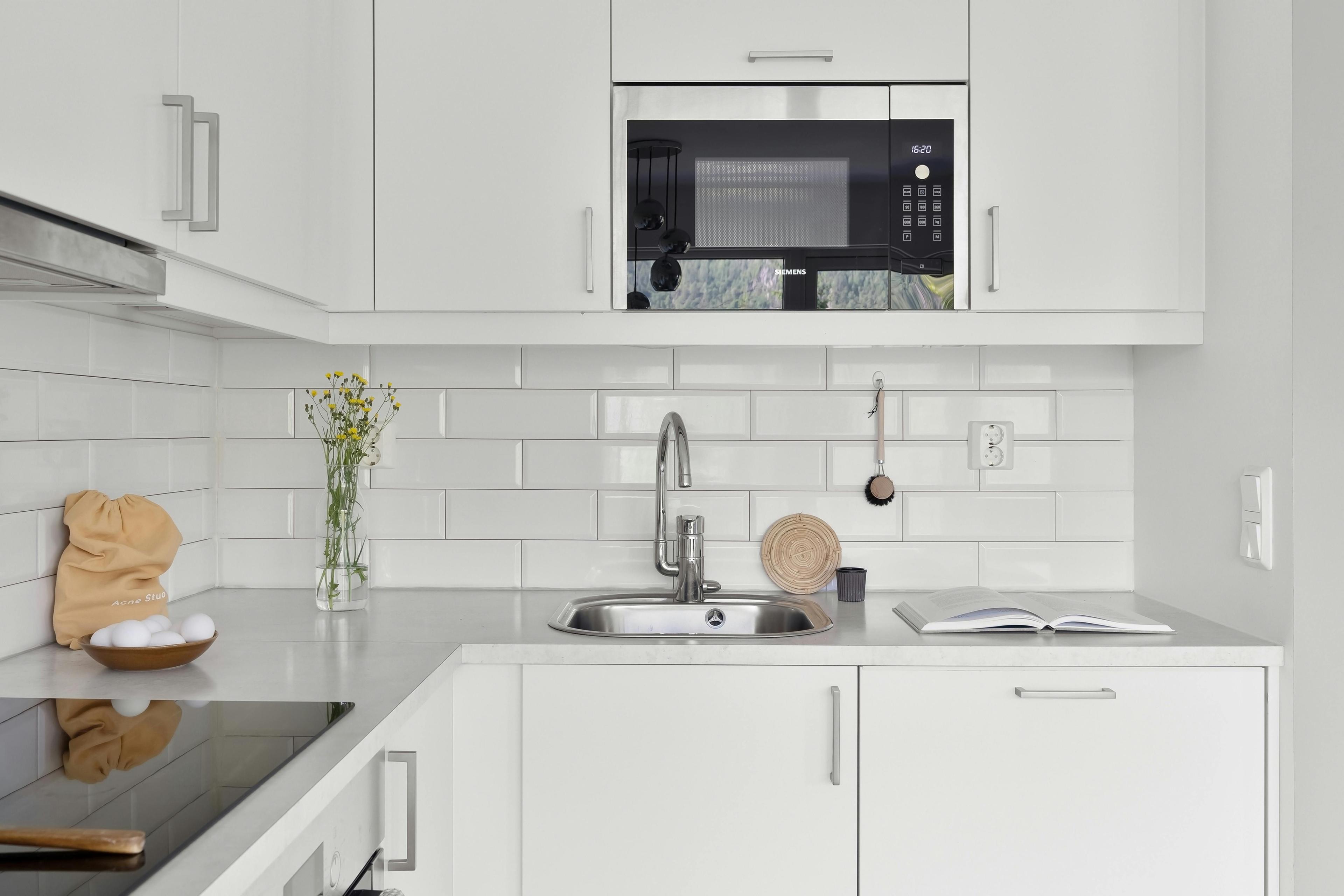 Moderne weiße Küchenzeile mit U-Bahn-Fliesen, Edelstahlspüle und eingebauter Mikrowelle, dekoriert mit Blumenvase und Küchenutensilien auf heller Arbeitsplatte.