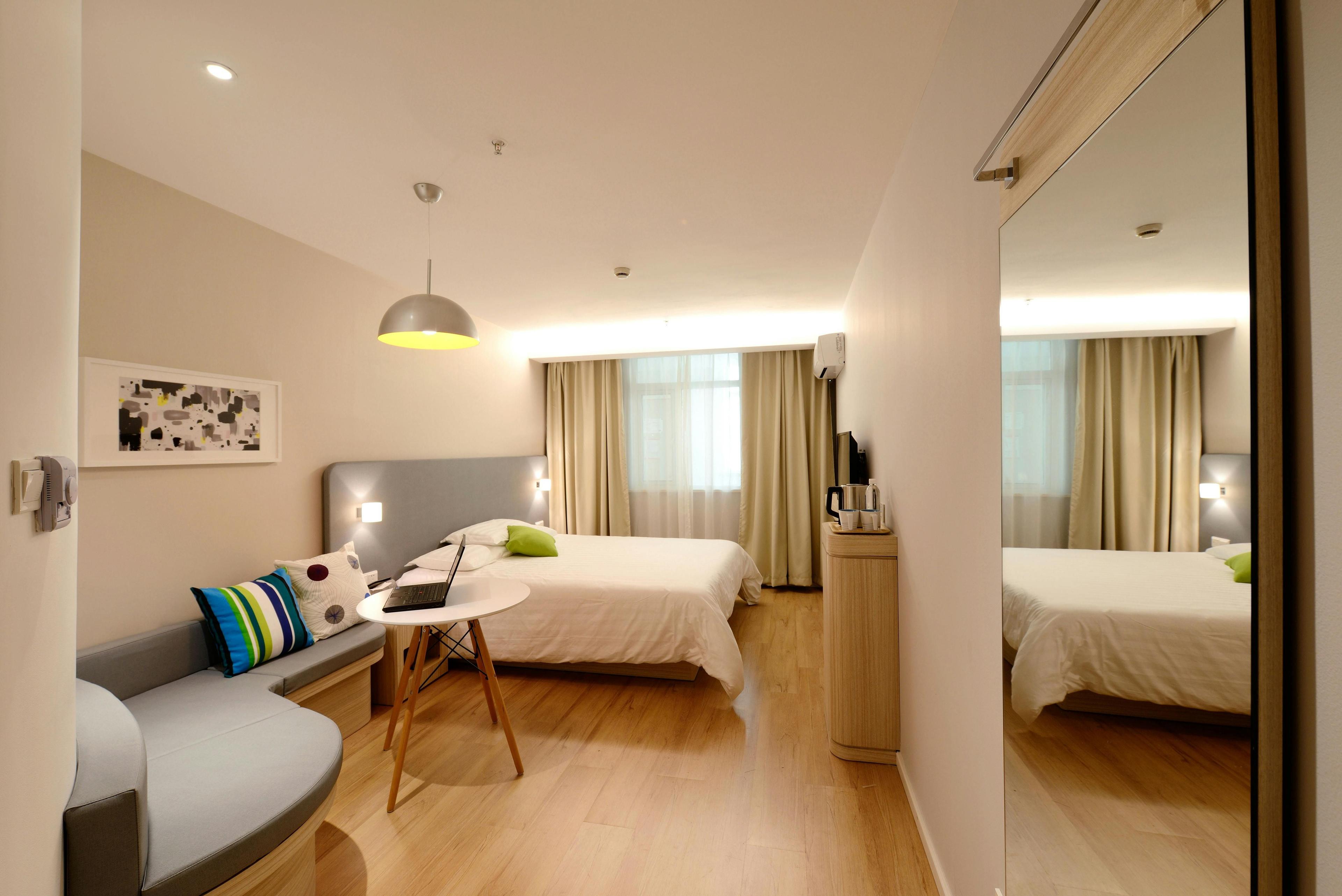 Helles Hotelzimmer mit Doppelbett, grauem Sofa, Spiegelschrank und rundem Arbeitstisch, Holzboden und großes Fenster mit beigen Vorhängen. Helles Hotelzimmer mit Doppelbett, grauem Sofa, Spiegelschrank und rundem Arbeitstisch, Holzboden und großes Fenster mit beigen Vorhängen.